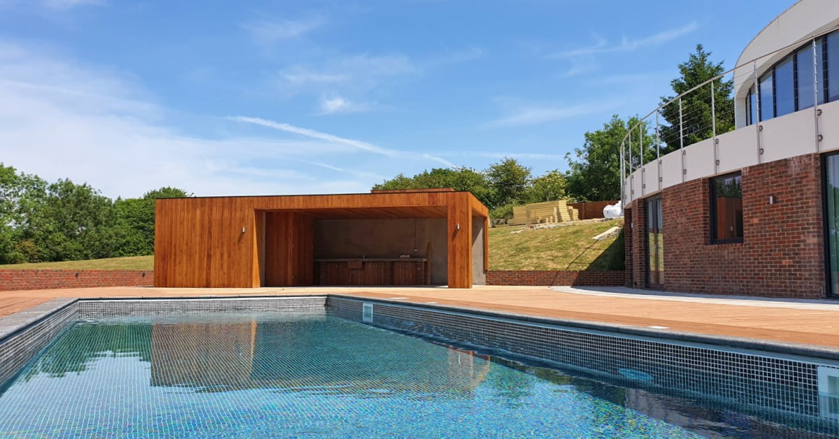 A modern outdoor pool with clear blue water is bordered by a sleek stone deck. On one side sits a contemporary wooden cabana with an open kitchenette, while the other side features a curved, glass-fronted building. The backdrop includes lush greenery and a clear blue sky, ensuring luxury at any cost. - a room in the garden