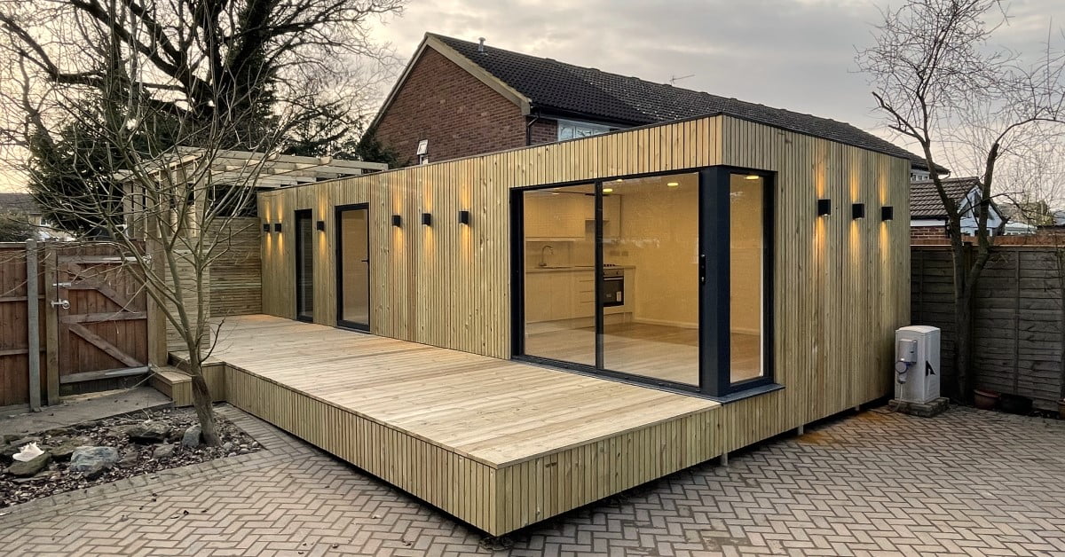 A cost-effective modern wooden outbuilding with large windows and black frames is situated in a backyard. It features a spacious wooden deck in front. Outdoor lights are mounted along the building's exterior, offering both style and function. The background includes a brick house, a wooden fence, and bare trees. - a room in the garden