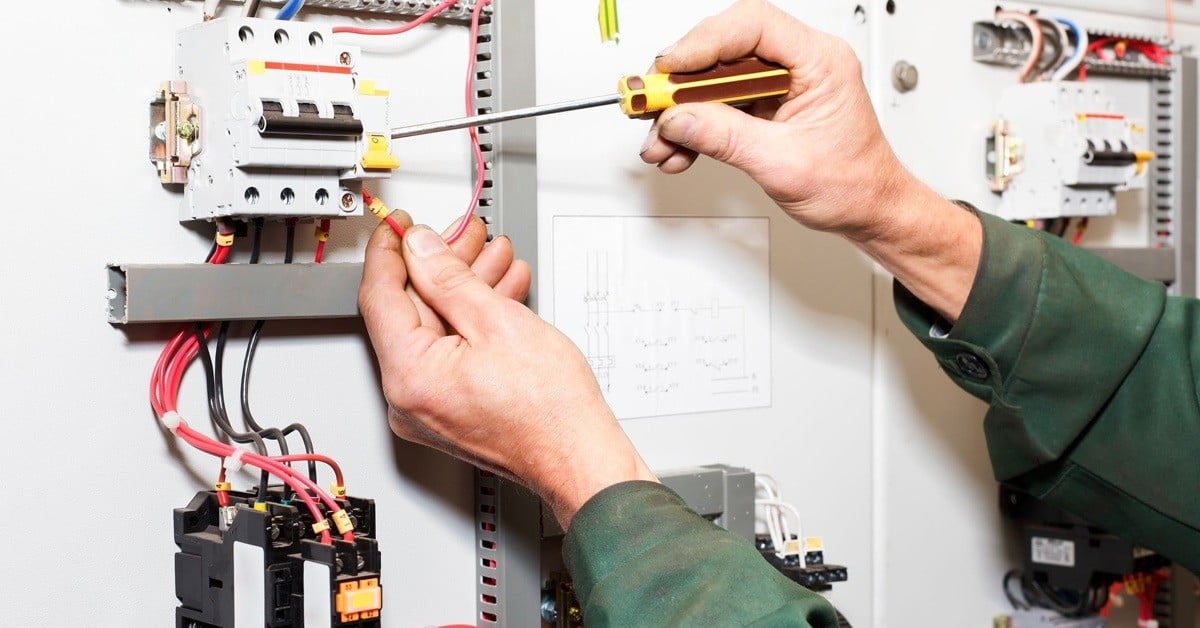 Close-up of a person using a screwdriver to adjust an electrical component in a control panel, highlighting the attention to detail that can reduce potential cost. The person's hands, wearing a green long-sleeve shirt, are visible along with various wires and electrical elements inside the open panel. - a room in the garden