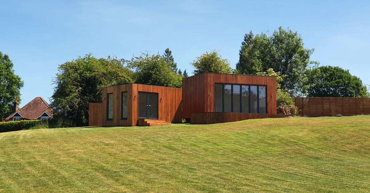 The image shows two modern, rectangular wooden houses with large glass windows set on a slightly sloped, lush green lawn. Tall trees and a traditional house with a steeply pitched roof are in the background. This picturesque setting accentuates the potential cost of luxurious living spaces. The sky is clear and blue. - a room in the garden