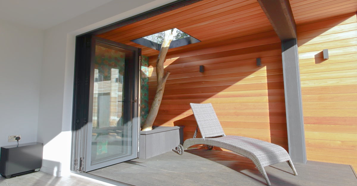 A cozy indoor-outdoor space featuring a wooden ceiling and walls, with a skylight allowing sunlight to stream in. A wicker lounge chair sits near a potted tree, and large folding glass doors open to the exterior, offering an inviting ambiance without the high cost of extensive renovations. - a room in the garden