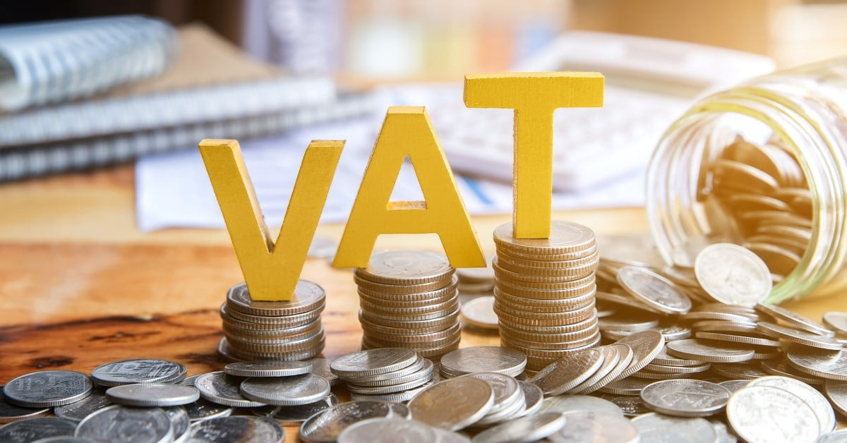 The image features the letters "VAT" standing on stacks of coins, symbolizing Value Added Tax. The background shows a blurred setting with documents, a calculator, and more coins spilling from a jar, highlighting the financial context and associated costs. - a room in the garden