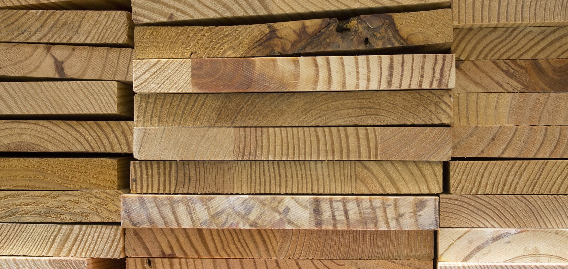 A stack of rectangular wooden planks neatly arranged in horizontal rows. The wood has natural grain patterns and varied shades of brown, with some planks showing unique knots and textures. Despite their beauty, the cost remains reasonable, making these straight-edged planks a valuable choice. - a room in the garden