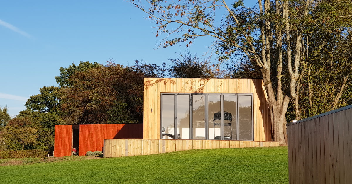 Modern wooden house with large windows situated on a grassy hill. The facade is minimalist with light wooden panels. Tall trees surround the property, and a bright blue sky is visible. Another smaller wooden structure with a red finish serves as a garden gym in the background. - a room in the garden