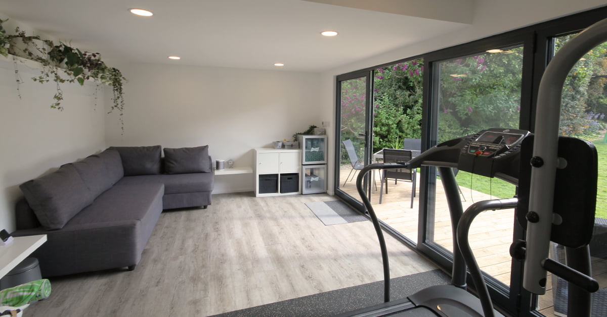 A modern garden gym with a treadmill facing sliding glass doors that open to a lush garden. There's a gray sectional sofa along the left wall, white shelves with storage cubes and decor on the right, and a light wood floor enhances the space's bright, airy feel. - a room in the garden