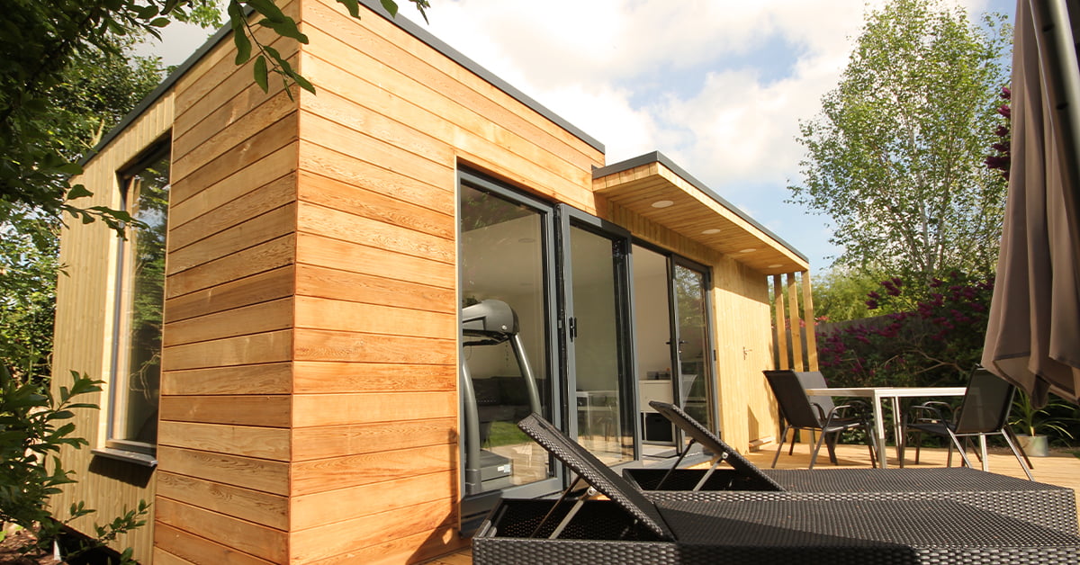 A modern wooden garden room with large sliding glass doors is nestled among lush greenery. This versatile space serves as a serene garden gym, with two black sun loungers on a wooden deck next to a small dining table and chairs under a patio umbrella. The sky is partly cloudy. - a room in the garden