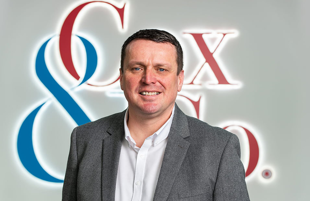 A man wearing a gray blazer over a white shirt is smiling at the camera. The background features the stylized company logo of Cox & Co., with an ampersand sign in blue and the text in red, reflecting their property management expertise. - a room in the garden