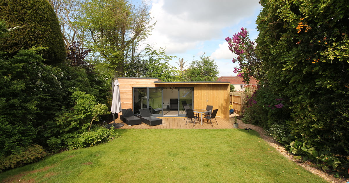 A modern wooden garden shed with large glass doors is surrounded by lush green trees and shrubs. The shed, constructed with SIP panels, sits on a wooden deck with outdoor furniture, including loungers and a small table. The foreground has a well-maintained grassy lawn. The sky is partly cloudy. - a room in the garden