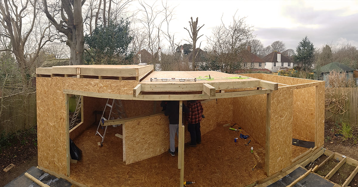 A panoramic view of a partially constructed building, likely a wooden shed or small house, with two people working inside. The structure's SIP panels and OSB walls are visible alongside the bare wooden frame. Tools and materials are scattered on the ground. Leafless trees surround the area. - a room in the garden