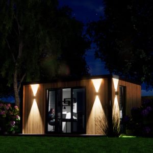 A small, modern wooden building is lit up at night by outdoor wall lights. The building has large glass doors, and a person is visible inside, sitting at a desk. The surrounding area features trees, shrubs, and grass, creating a peaceful nighttime scene. - a room in the garden