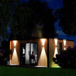 A modern, small wooden cabin is illuminated at night by outdoor sconces casting warm light. Through the sliding glass doors, a person can be seen sitting at a desk inside, working or reading. The cabin is surrounded by trees and shrubs, creating a tranquil atmosphere. - a room in the garden