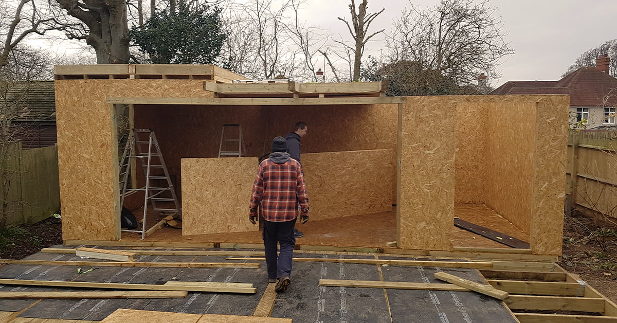 Two people are assembling a large wooden shed in a backyard. One person, wearing a plaid jacket, walks towards the shed while another inside lifts a SIP panel. Unfinished framing and other construction materials are scattered around the area. - a room in the garden