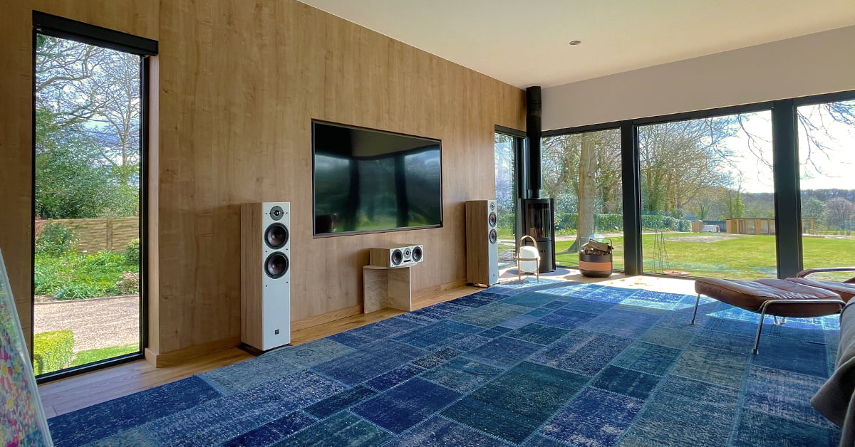 A spacious living room with a modern design features a large flat-screen TV mounted on a wooden wall. The floor is covered with a blue patterned carpet. Tall windows and a glass door offer a view of the green lawn and trees outside, flooding the property with natural light. - a room in the garden