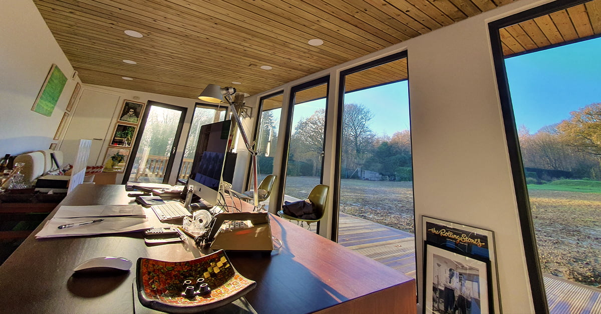 A modern home office with a wooden ceiling, large windows, and a view of a grassy outdoor area with trees. This stunning property features a desk equipped with a computer, keyboard, mouse, lamp, and decorative bowl. A framed picture and various office items adorn the desk and shelves. - a room in the garden