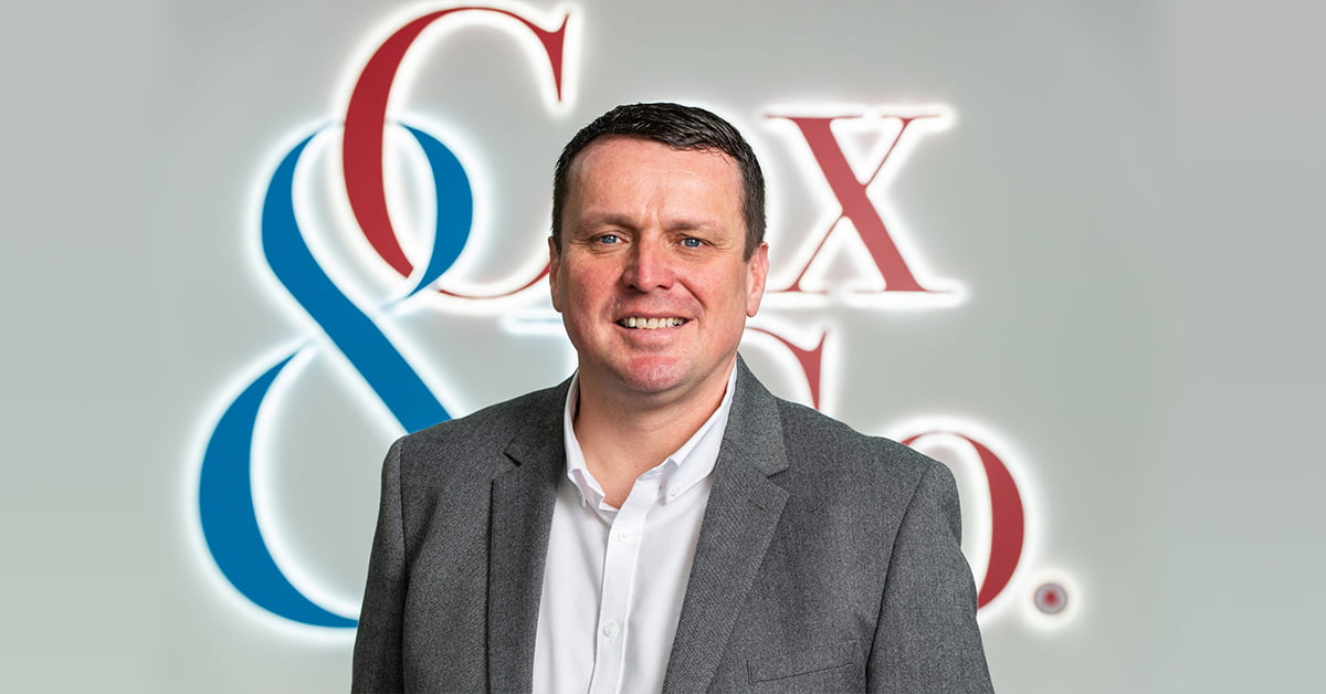 A man in a gray blazer and white shirt is smiling at the camera. Behind him, a blurred company logo featuring the text "Cox & Co." in blue and red hues hints at their property services. The background is light gray. - a room in the garden