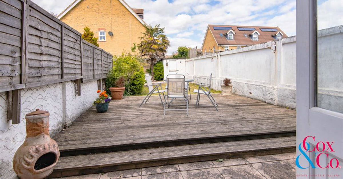 A charming property featuring a backyard with a wooden deck adorned with a metal table and chairs set. A potted plant rests beside the table, all enclosed by a white fence and wooden panels. In the background stands a brick house amidst lush greenery, under a partly cloudy sky. - a room in the garden