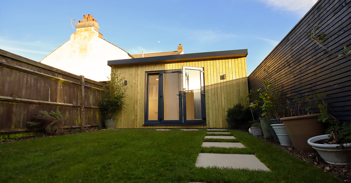 A small modern garden shed with wooden cladding and dark-framed glass doors sits on a neatly manicured lawn. Stone stepping stones lead from the lawn to the shed. The property is enclosed by wooden fences, and potted plants decorate the area. The sky is clear. - a room in the garden