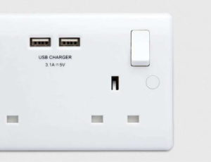 A white electrical wall socket featuring two USB charging ports (3.1A = 5V) positioned at the top. To the right of the USB ports, there is a switch, and below it, various plug socket openings. The wall background is light grey. - a room in the garden