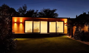 A small, modern house with large windows illuminated from the inside at dusk. The wooden exterior glows with warm lighting, and the house is surrounded by trees and a neatly trimmed lawn. An exercise bike is visible through the window. - a room in the garden