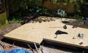 A garden building project is underway with a flat wooden platform under construction in a backyard area. Various tools and items like a broom, fabric, and a bottle are scattered on the surface. The ground around the platform shows disturbed soil and construction materials. Trees and a wooden fence surround the area. - a room in the garden