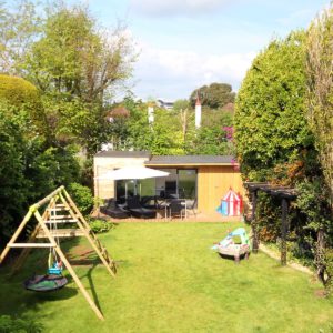 A lush backyard features a wooden play structure with swings on the left, a grassy lawn in the center, and a patio area with outdoor furniture and umbrellas in the background. A small play tent and various toys are scattered around the lawn. - a room in the garden