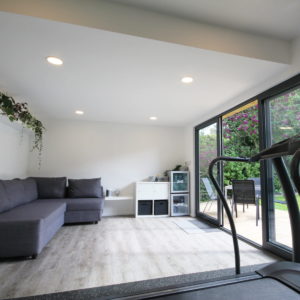 A modern home gym features a gray sectional sofa, a treadmill, a white cabinet, and a wall-mounted shelf with plants. Large glass sliding doors on the right allow natural light and open to a patio with a small dining set and lush greenery. - a room in the garden