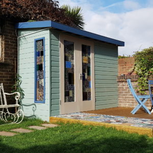 A small, light blue wooden garden shed with a flat dark blue roof featuring colorful stained glass panels on the doors and window. The shed is situated in a grassy yard, adjacent to a brick wall with greenery, and a white metal bench and blue table visible to the right. - a room in the garden