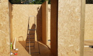 A partially constructed wooden garden building with oriented strand board (OSB) walls is shown. A metal step ladder stands in the center, surrounded by some construction materials and tools lying on the floor. Sunlight casts shadows inside the structure. - a room in the garden
