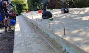 A close-up of a garden building construction site where workers are installing an insulated floor. Several wooden boards are laid out with screws partially drilled in. Tools, including a drill, are scattered on the floor. One worker is kneeling in the background using a tool. - a room in the garden