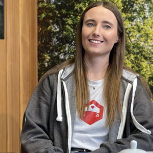A woman with long brown hair smiles while sitting indoors. She is wearing a white T-shirt with a red logo, a gray zip-up hoodie, and is in front of a wooden wall with a window revealing greenery outside. The lighting is soft, emphasizing her joyful expression. - a room in the garden