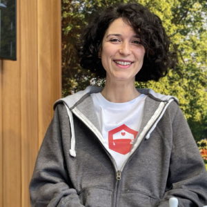 A person with curly dark hair, wearing a grey hoodie and a white t-shirt with a red heart symbol, is smiling while sitting indoors. The background features wooden panels and a lush green outdoor scene. - a room in the garden