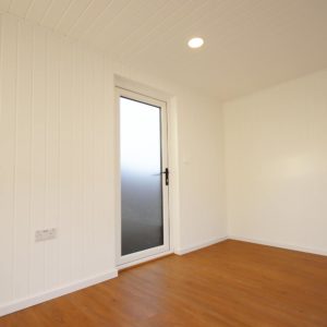 A small, empty room with white paneled walls and a wooden floor. There is a closed frosted glass door with a black handle on the left wall, leading outside. A single round ceiling light is on, illuminating the space. - a room in the garden