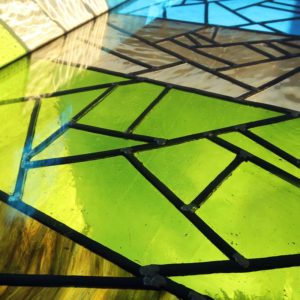 A close-up photo of a stained glass window, featuring geometric shapes with black outlines. The glass sections are in shades of green, blue, and earthy brown colors, with light shining through, creating a mix of bright and subtle hues. - a room in the garden