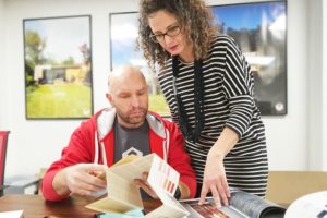 A man and a woman are collaborating at a table. The man, wearing a red hoodie, is reviewing a brochure, while the woman with curly hair, in a black and white striped dress, is pointing to a book on the table. They appear to be discussing design plans. - a room in the garden