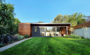 A modern single-story house with wooden paneling, large glass sliding doors, and a well-manicured green lawn in the foreground. The backyard features various plants and trees along the perimeter, and the clear blue sky is visible above. - a room in the garden