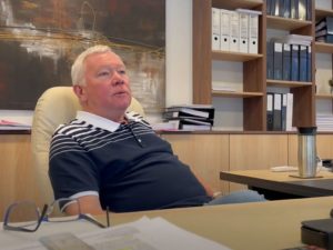 A man with light hair and a striped polo shirt is sitting at a desk in an office. Behind him are shelves with binders and stacks of papers. A stainless steel travel mug is on the desk, and a pair of glasses rests on scattered papers in the foreground. - a room in the garden