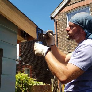 A person wearing a blue headscarf, gloves, and a light purple shirt is painting the edge of a wooden structure using a brush. The person focuses on applying paint to the wood. Brick buildings and a clear blue sky are visible in the background. - a room in the garden