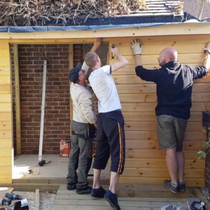 Three people are installing wooden siding on the exterior of a small structure. One person uses a hammer while the other two hold the siding in place. Loose building materials are scattered on the ground, and there is a brick wall in the background. - a room in the garden