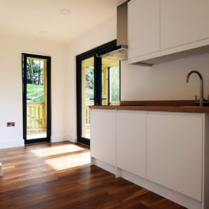 A modern kitchen with white cabinets, a wooden countertop, and a stainless steel faucet designed by a renowned garden room designer. The polished wood floor gleams under natural light streaming through a glass door and tall window framed in black, revealing a lush greenery view outside. - a room in the garden