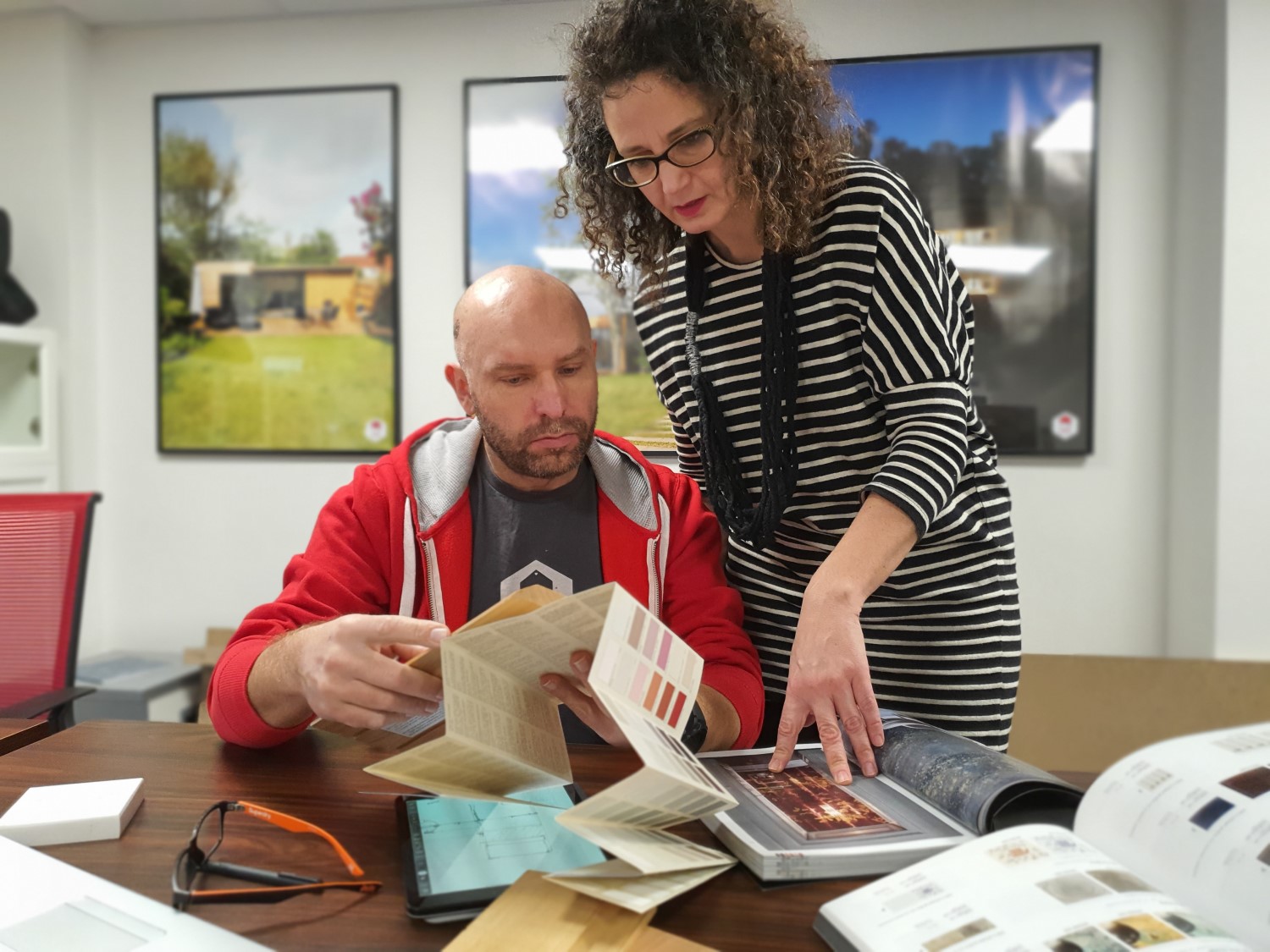 A man and a woman, likely designers, are reviewing design materials at a table covered with samples, catalogs, and a tablet. The man, in a red hoodie, holds some samples while the woman in a striped dress stands beside him, pointing at a catalog possibly for their garden house project. - a room in the garden