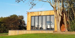 A modern, small wooden building with large glass doors stands on a slightly elevated wooden deck surrounded by green grass. A leafless tree is in the foreground, and bushes are visible in the background under a clear blue sky. - a room in the garden