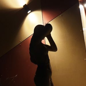 A silhouette of a person standing against a two-toned wall, illuminated by overhead lighting. The person appears to be adjusting or holding something near their face, with their back slightly arched, creating an artistic and dramatic shadow effect. - a room in the garden