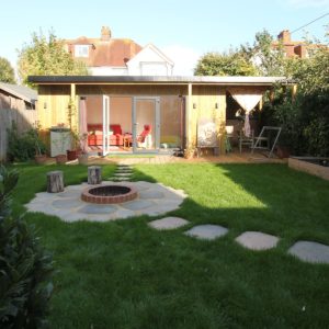 A backyard features a green lawn with stone pavers leading to a stone fire pit surrounded by tree stump stools. At the back, a wooden building with large glass doors opens to a patio resembling an outdoor gallery, adorned with potted plants, a hammock, and a swing seat. Trees and houses are visible beyond. - a room in the garden