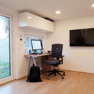 A modern home office with a wooden desk holding a computer monitor, a keyboard, a tablet, and other accessories. A black office chair is placed in front of the desk. A large window on the left and a mounted TV on the right wall are visible. A black backpack rests on the floor. - a room in the garden