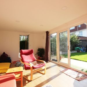 A bright, sunlit room with large glass doors opening to a garden resembles an art gallery. The room features a wooden floor, a red armchair with a footrest, a yellow sofa, and a small wooden table. A potted plant near the glass doors and a small rug on the floor complete the inviting space. - a room in the garden