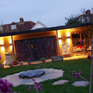 A cozy backyard scene at dusk featuring a wooden house with large glass doors, warmly lit from the outside. There is a fire pit in the foreground surrounded by a patio that feels like an outdoor gallery. Decorative plants and a statue with a Buddha face adorn the area. - a room in the garden