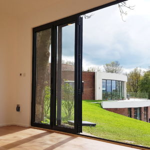 A modern room with large glass sliding doors open to a scenic view of a green lawn and a contemporary building with large windows. The sky is partly cloudy, and trees surround the buildings. The interior has light wood flooring and white walls. - a room in the garden