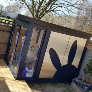 A small, modern shed with dark blue panels and large windows is in a garden. A stylized bunny shape decorates the front panels. The shed is surrounded by a wooden fence and trees, and a wooden deck extends in front of the shed. Bright, sunny day. - a room in the garden