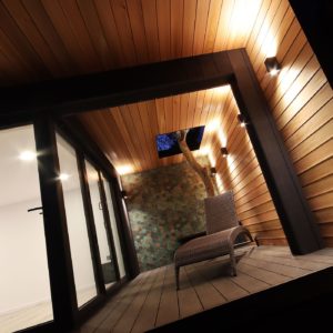 A modern, warmly lit wooden patio features a lounge chair on a wooden deck. The ceiling and walls are lined with wood panels, with recessed lighting on the ceiling. A partial wall with decorative tiles showcases a built-in tree, creating a serene, cozy ambiance. - a room in the garden