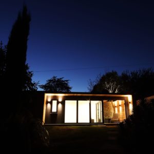 Modern single-story house with floor-to-ceiling windows aglow at night. The minimalistic design features a flat roof and a blend of warm and cool exterior lighting. Designed by a talented garden room designer, the home is surrounded by silhouetted trees and foliage, creating a serene and inviting scene. - a room in the garden